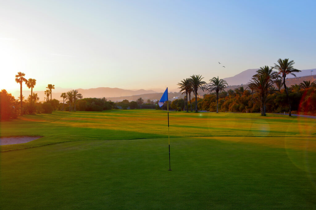 A hole with a blue flag at Almerimar Golf Course at sunset