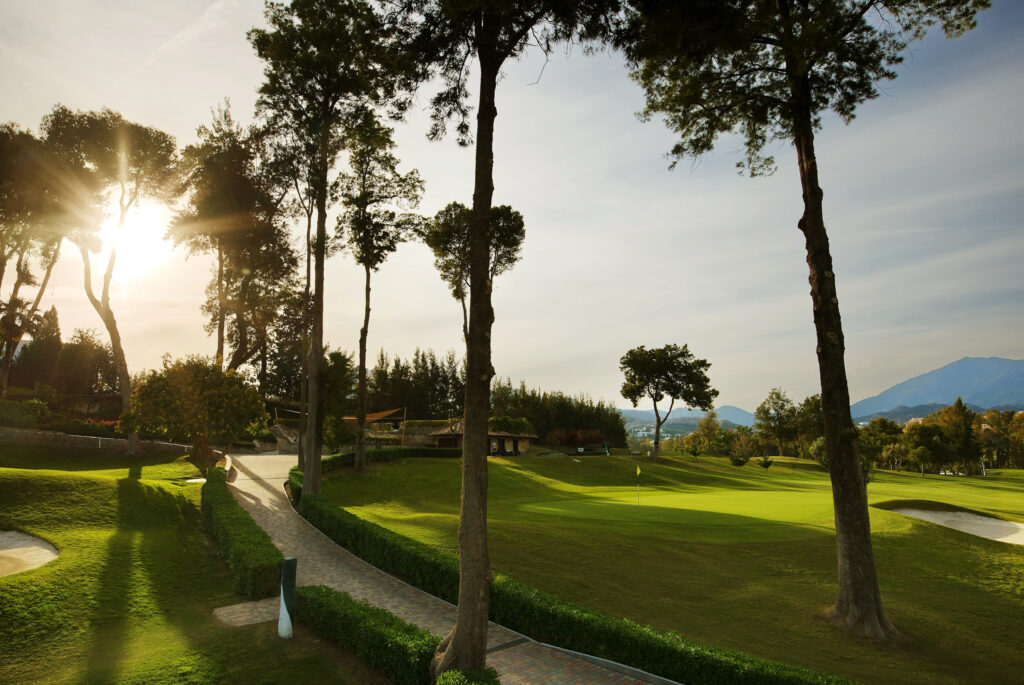 Path leading past a hole with yellow flag with trees around at Atalaya - Old Golf Course