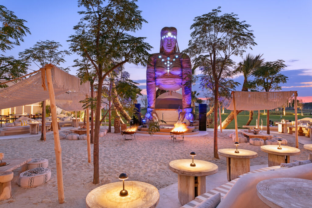 One of the many bar areas that has a man made beach bar style. The bar has white sand and fire pits surrounded by comfortable seating, with a stage for events and a large statue engulfing the background