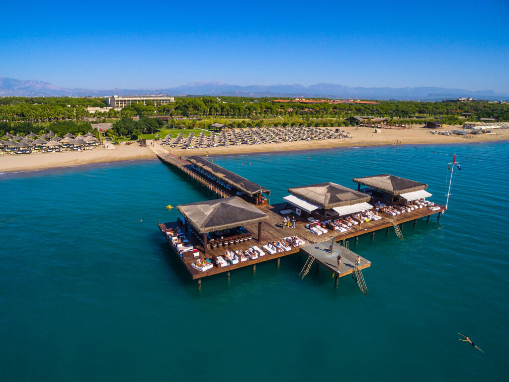 A panoramic view of the belek landscape and the Gloria Serenity Resort. The Sea & Beach are in the foreground with the luxurious pier that extends out into the ocean, and the hotel and gardens make up the background with the bright blue sky above.