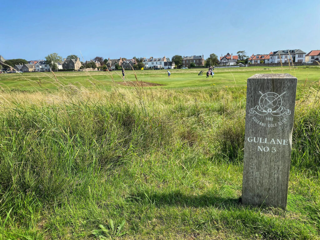 Gullane Golf Club – No. 3 Course wooden post with people playing golf in background