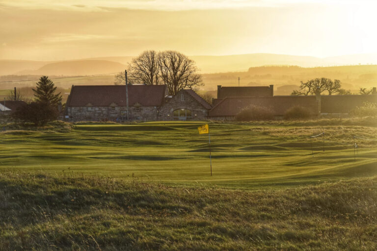 Hole with yellow flag at Gullane Golf Club – No. 3 Course with building in background at sunset