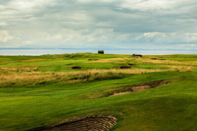 Fairway at Kilspindie Golf Club leading to hole with red flag and bunkers at Kilspindie Golf Club with ocean in background