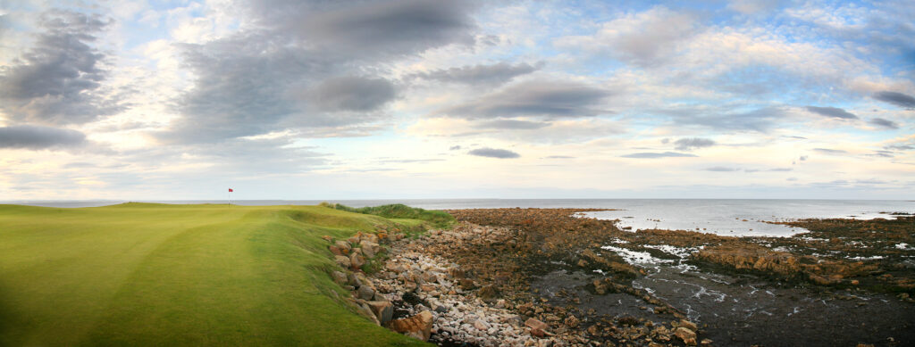 Panoramic view of a hole with a red flag at Kingsbarns Golf Links and the beach