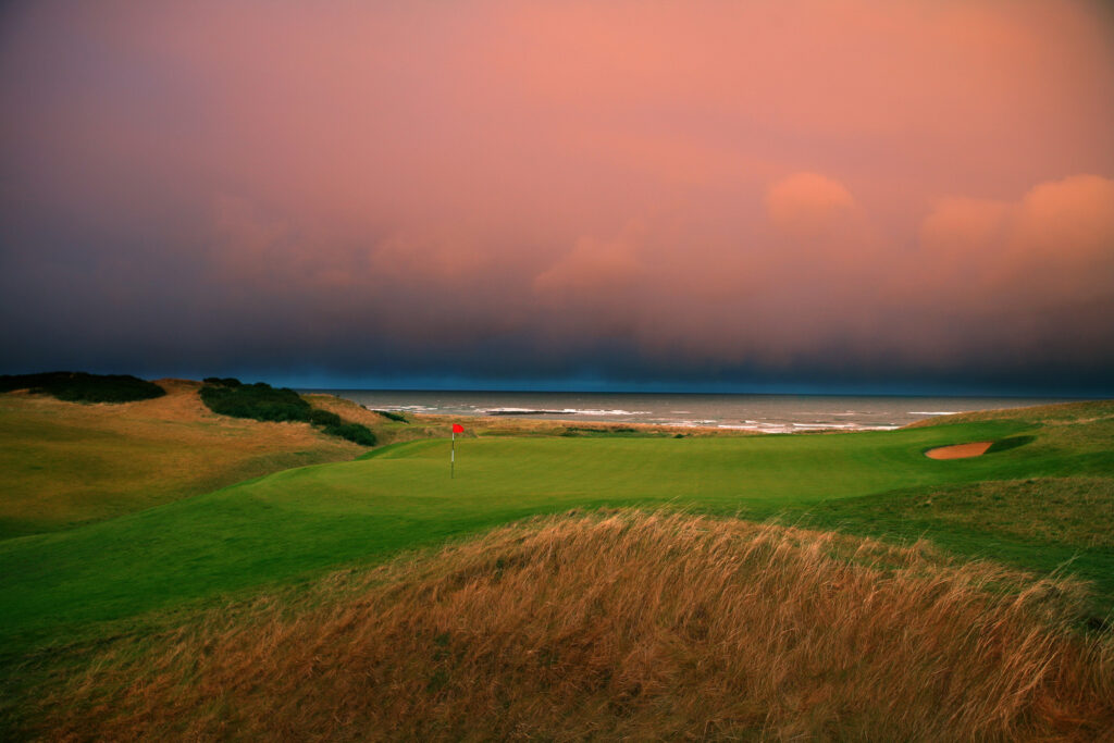 Hole with red flag at Kingsbarns Golf Links with ocean in background at sunset