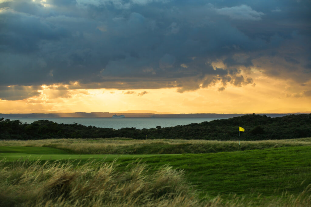 Hole with yellow flag at Muirfield with ocean in background