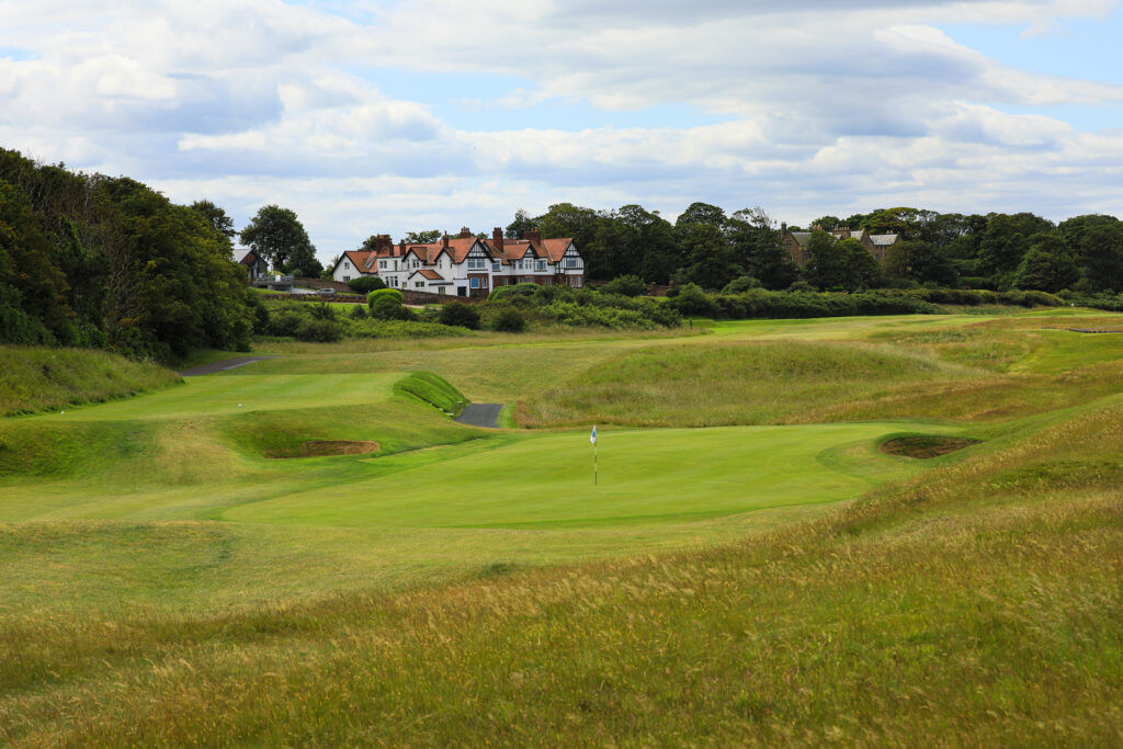 Hole with white flag at North Berwick Golf Club with building in distance and trees around