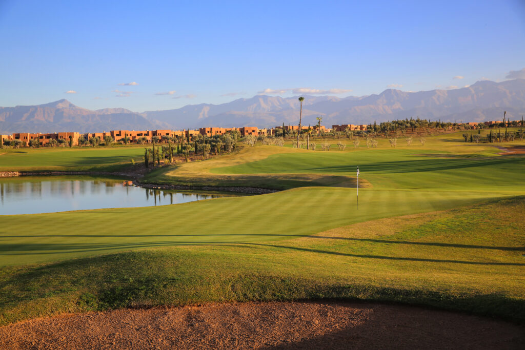 Hole with bunker and lake at Palm Golf Ourika with trees, buidings and mountains in background