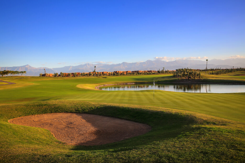 Hole with bunker and lake at Palm Golf Ourika with buildings and mountains in distance