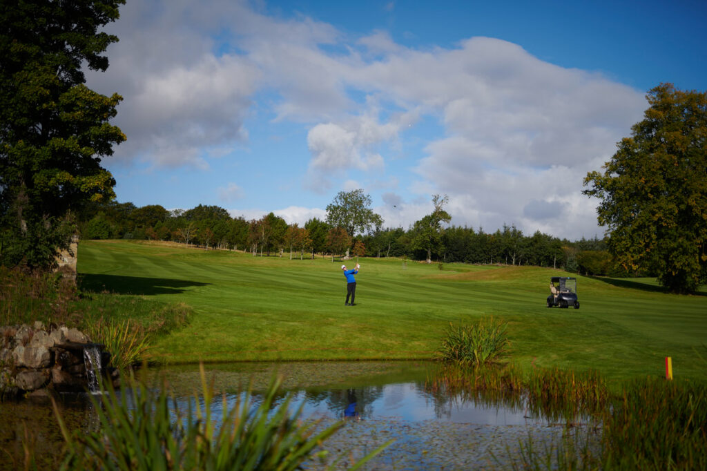 Person playing golf at Meldrum House Golf Club with lake in foreground and trees in background