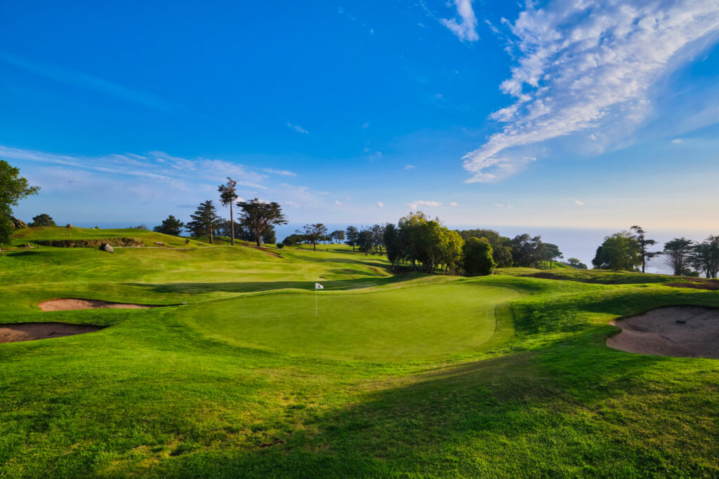 Hole with white flag with trees in background at Aerial view of a hole overlooking the ocean at Palheiro Golf Course