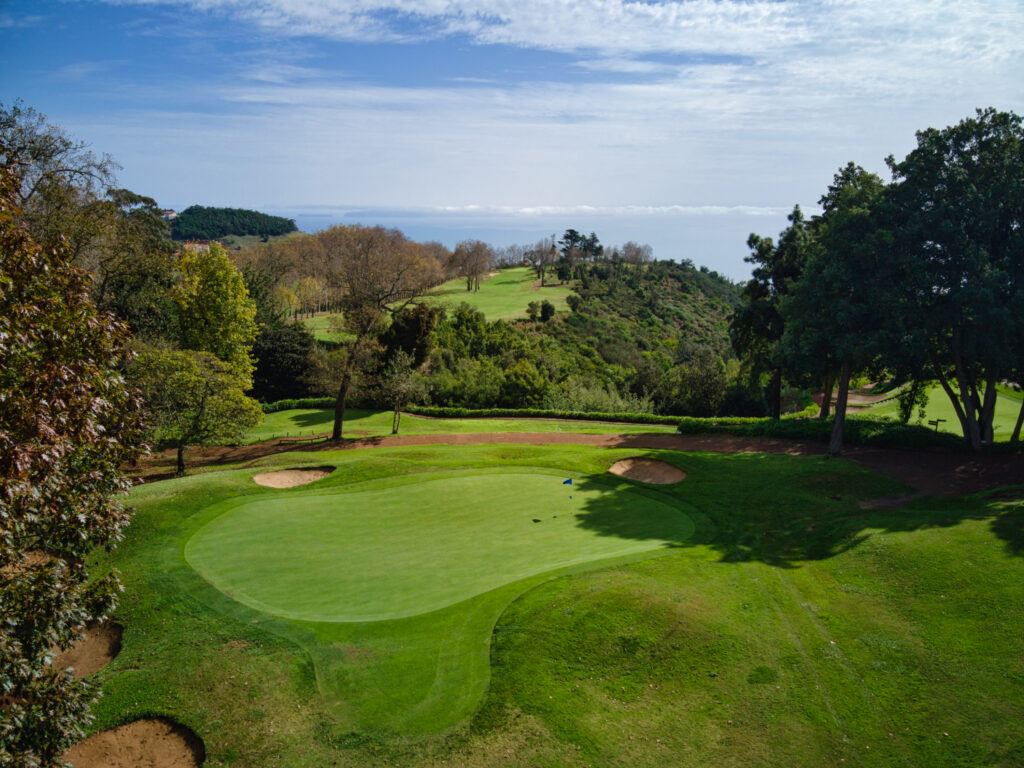 Hole with blue flag with bunkers and trees around at Aerial view of a hole overlooking the ocean at Palheiro Golf Course