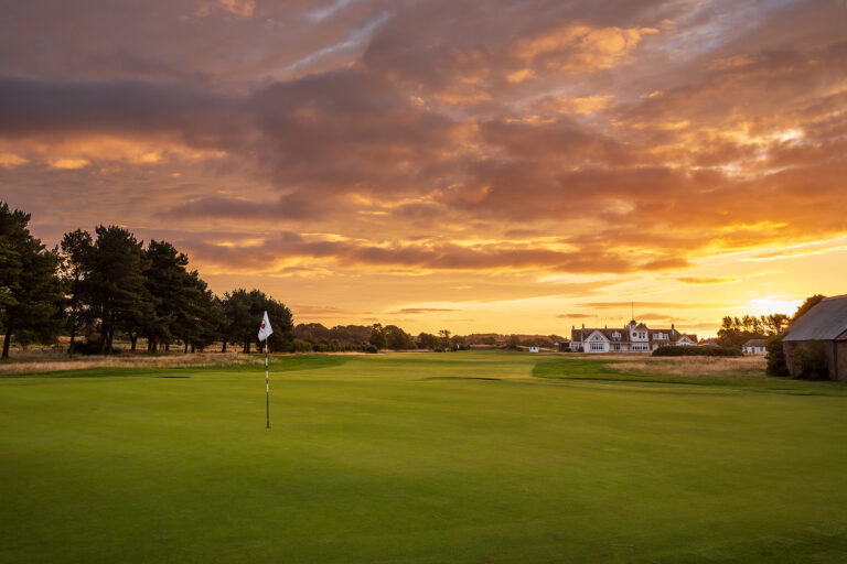 Hole with white flag at Panmure Golf Club with building in distance at sunset