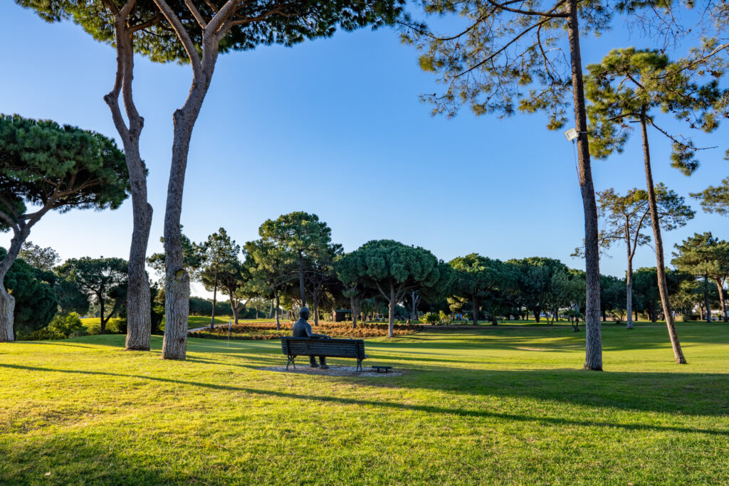 Statue of man sitting on a bench looking into the fairway with trees around at Vila Sol Golf Course