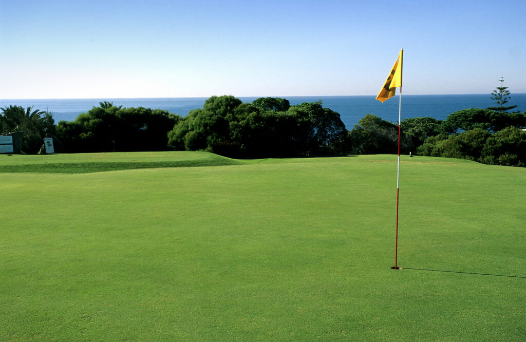 Hole with yellow flag with trees and the ocean in the background at Onyria Quinta da Marinha Golf Course