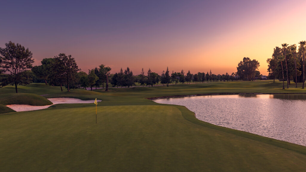 A hole with yellow flag with a lake and bunker and trees around at Real Club Sevilla Golf