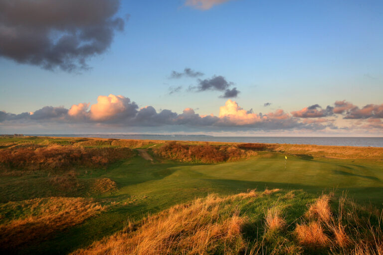 Hole with yellow flag at Royal Cinque Ports Golf Club with mounds around and ocean in background