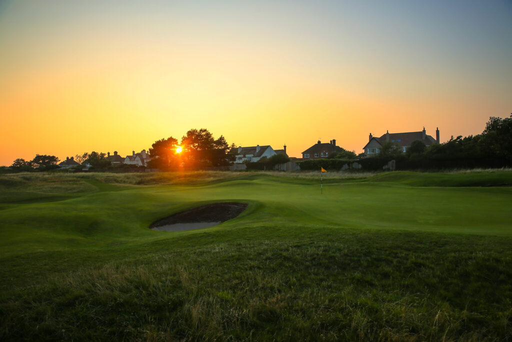 Hole with yellow flag with bunker at Royal Liverpool Golf Club with buildings in distance