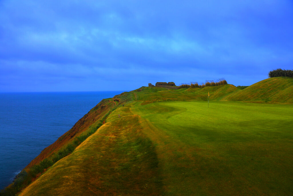 Hole with yellow flag at Old Head Links with ocean view and building in distance