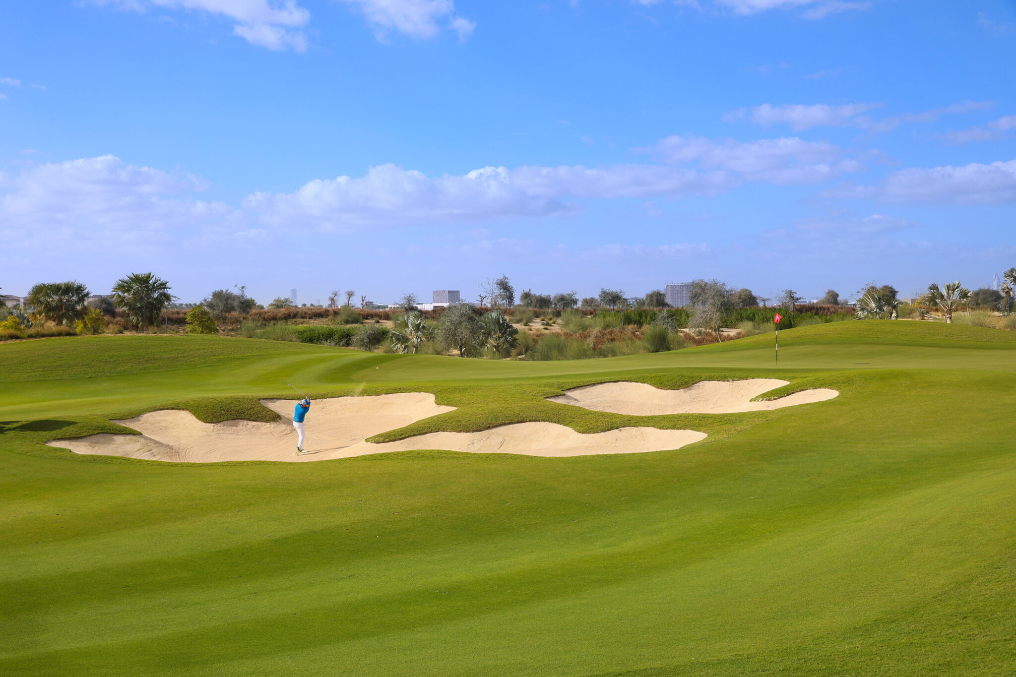 Person playing golf out of a bunker at Dubai Hills Golf Club with trees in background